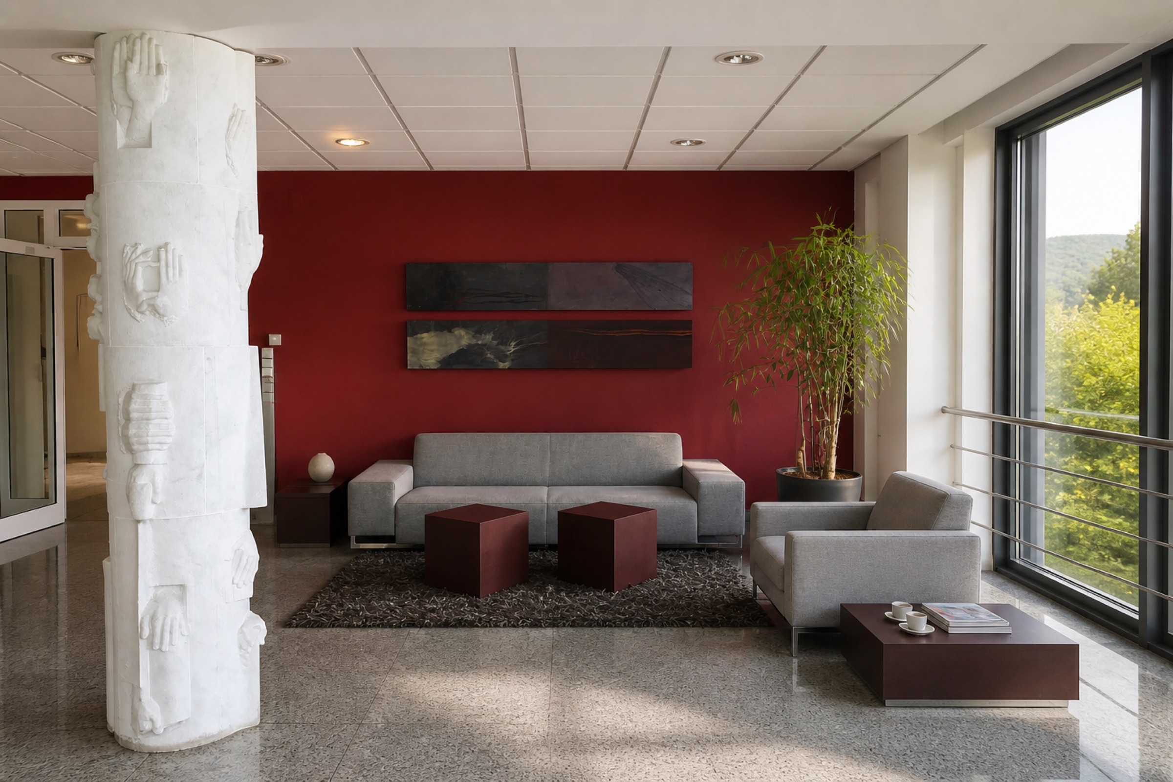 Offener Aufenthaltsbereich mit weinroter Wand, grauen Sofas und bodentiefer Fensterfront ins Grüne